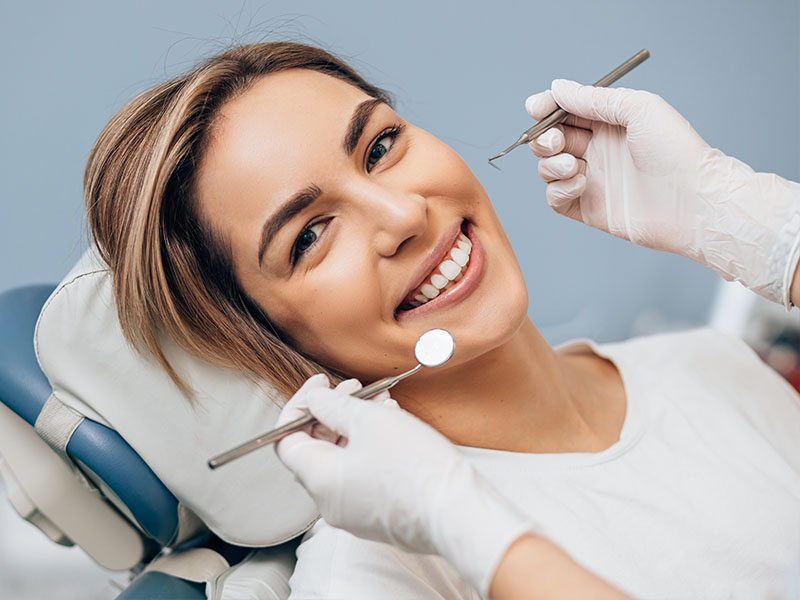 A woman is sitting in a dental chair, smiling, with her teeth being worked on by a dentist.