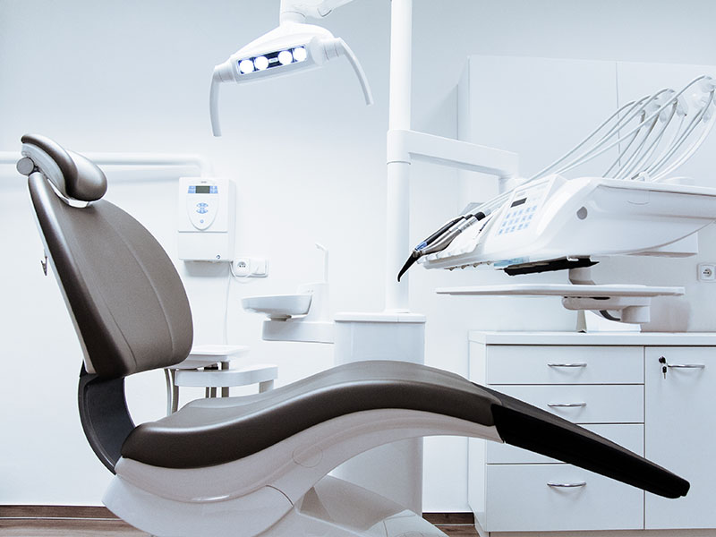 The image shows a dental office interior with a modern dental chair and equipment, including a dental light, in front of a large window.