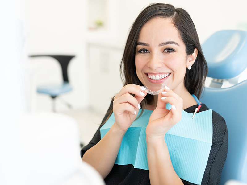 The image shows a person sitting in a dental chair, smiling at the camera while holding up a toothbrush. They are wearing a black top and appear to be in a dental office setting.