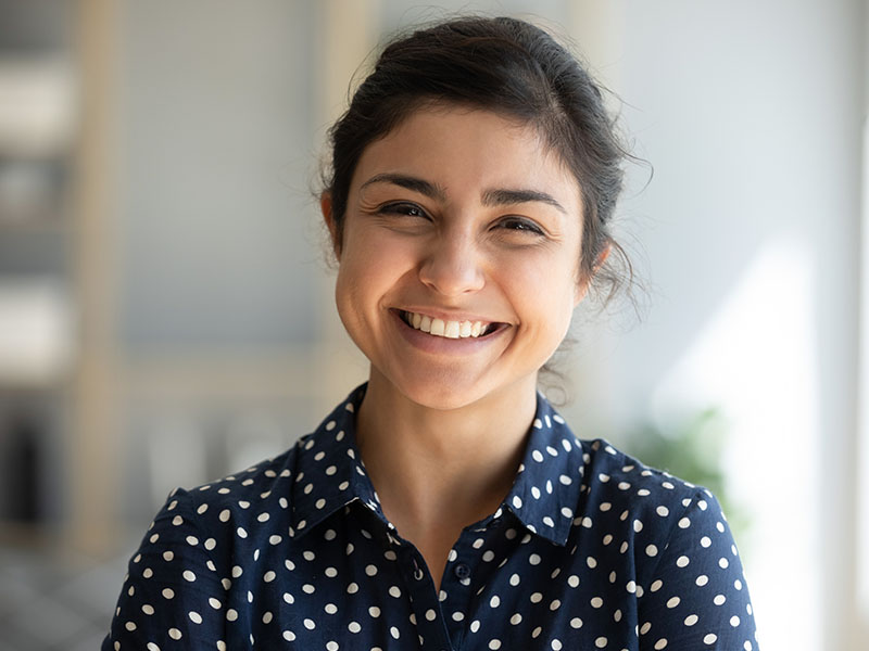 A young woman with a smile, wearing a polka dot blouse and a dark top, stands in an office setting.
