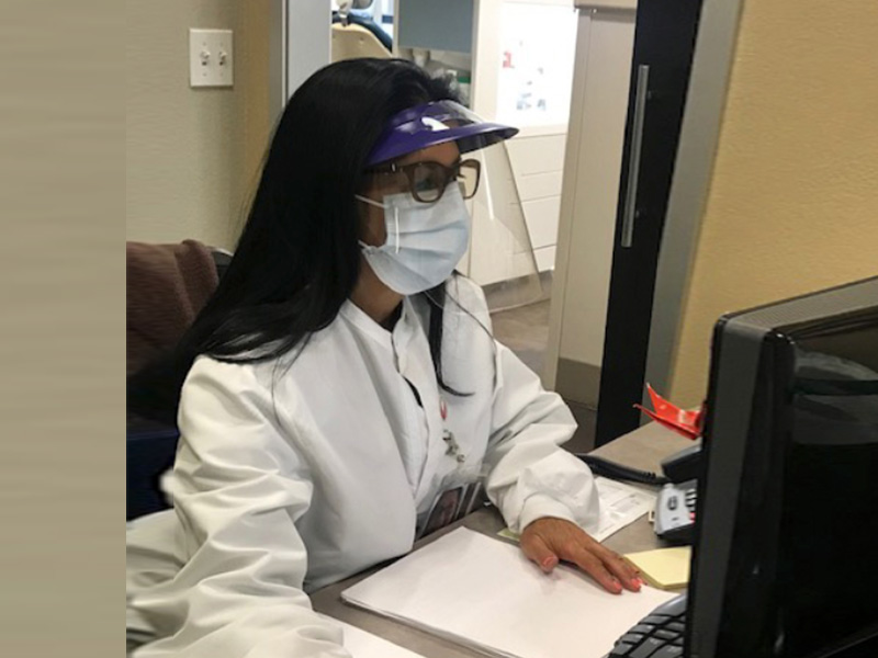 The image shows a person wearing glasses, a face mask, and a white lab coat sitting at a desk with a computer monitor in front of them.