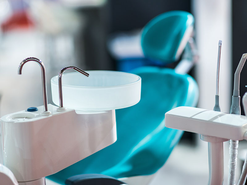 The image shows a modern dental office setting with a blue dental chair, a white dental machine, and various dental tools.