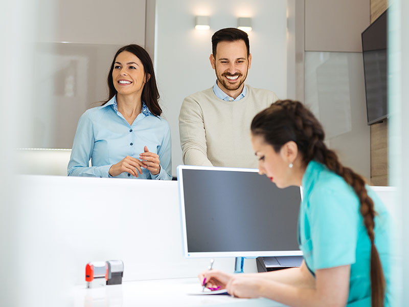 A professional setting with a man and woman in business attire, standing behind a desk, smiling at a female receptionist seated across from them.